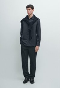 Donkergrijs overhemd van gladde stof, gecombineerd met loszittende donkere broek. Een zwarte trui ligt over de schouders gedrapeerd. Zwarte schoenen.