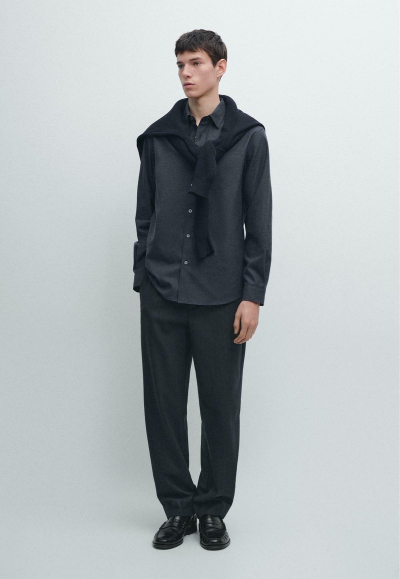 Donkergrijs overhemd van gladde stof, gecombineerd met loszittende donkere broek. Een zwarte trui ligt over de schouders gedrapeerd. Zwarte schoenen.