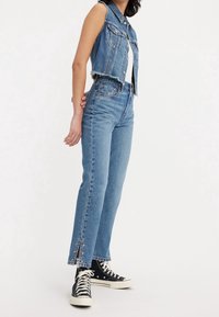 Mujer que lleva unos jeans de tiro alto en azul claro con cortes en los tobillos y detalles de estrellas doradas, combinados con una chaqueta vaquera sin mangas y zapatillas negras.