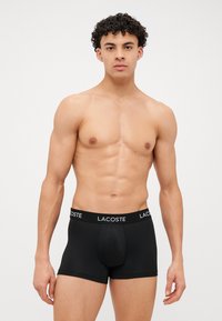 Črne bokserice z elastičnim pasom, na katerem je bel napis "LACOSTE". Gladek material, prilegan kroj in dolžina do sredine stegna.