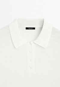 Valkoinen neulottu polo-paita, jossa on ribbauspääntie, kolme napinläpeä ja hienovarainen tekstuuri kankaan läpi. Brändilogo näkyvissä.