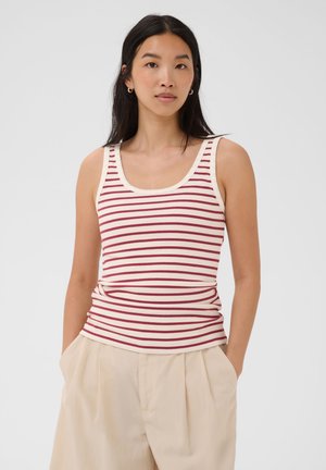 Femme portant un débardeur crème sans manches avec des rayures horizontales rouges et un pantalon beige taille haute, debout les mains dans les poches.
