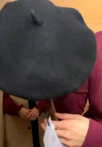 Personne portant un grand béret noir et un haut bordeaux tenant un objet blanc, avec une autre personne en manteau beige et manches rouges en arrière-plan.