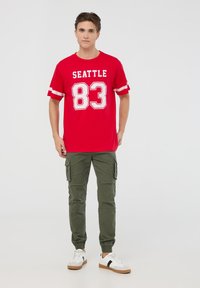 T-shirt di cotone rossa con la scritta "SEATTLE 83" stampata in bianco, con maniche corte a righe. Abbinata a pantaloni cargo verdi oliva e sneakers bianche.