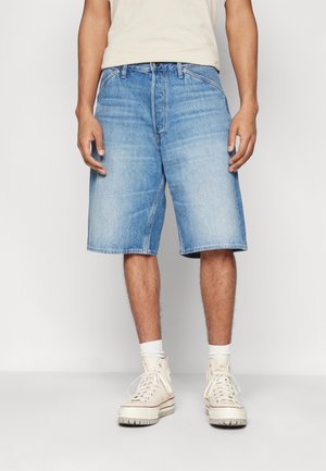 Homme portant un short en jean bleu clair, des chaussettes blanches et des baskets montantes écru, debout sur un fond clair uni.