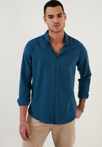 Camisa abotonada en azul oscuro, hecha de una tela suave. Presenta un collar puntiagudo, mangas largas remangadas y botones en contraste en la parte delantera.