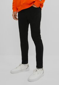 Černé skinny jeans vyrobené z denimu, spárované s bílými teniskami a oranžovou mikinou. Oblečení má výrazný volný styl a upnutý tvar.