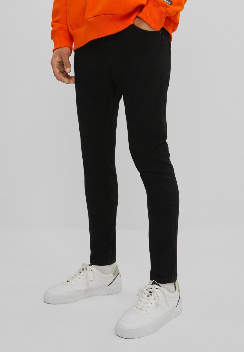 Černé skinny jeans vyrobené z denimu, spárované s bílými teniskami a oranžovou mikinou. Oblečení má výrazný volný styl a upnutý tvar.