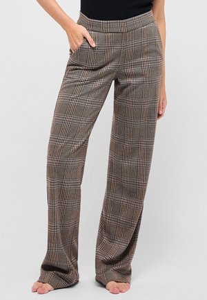 Kobieta w wysokich, szerokich spodniach w kratę, z rękami w kieszeniach, stojąca boso na jednolitym tle.