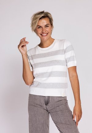 Smiling woman with short blonde hair wearing a white and gray striped short-sleeve shirt and gray pants, posing against light background.