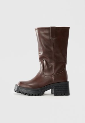 Brown mid-calf leather boot with thick black lug sole and block heel, featuring pull tab and stitched detailing.