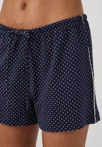 Tamsiai mėlynos polka dot šortai, pagaminti iš minkštos medžiagos, su elastinga juosmens juosta ir raišteliu, bei balta apdaila šoninėse pusėse.