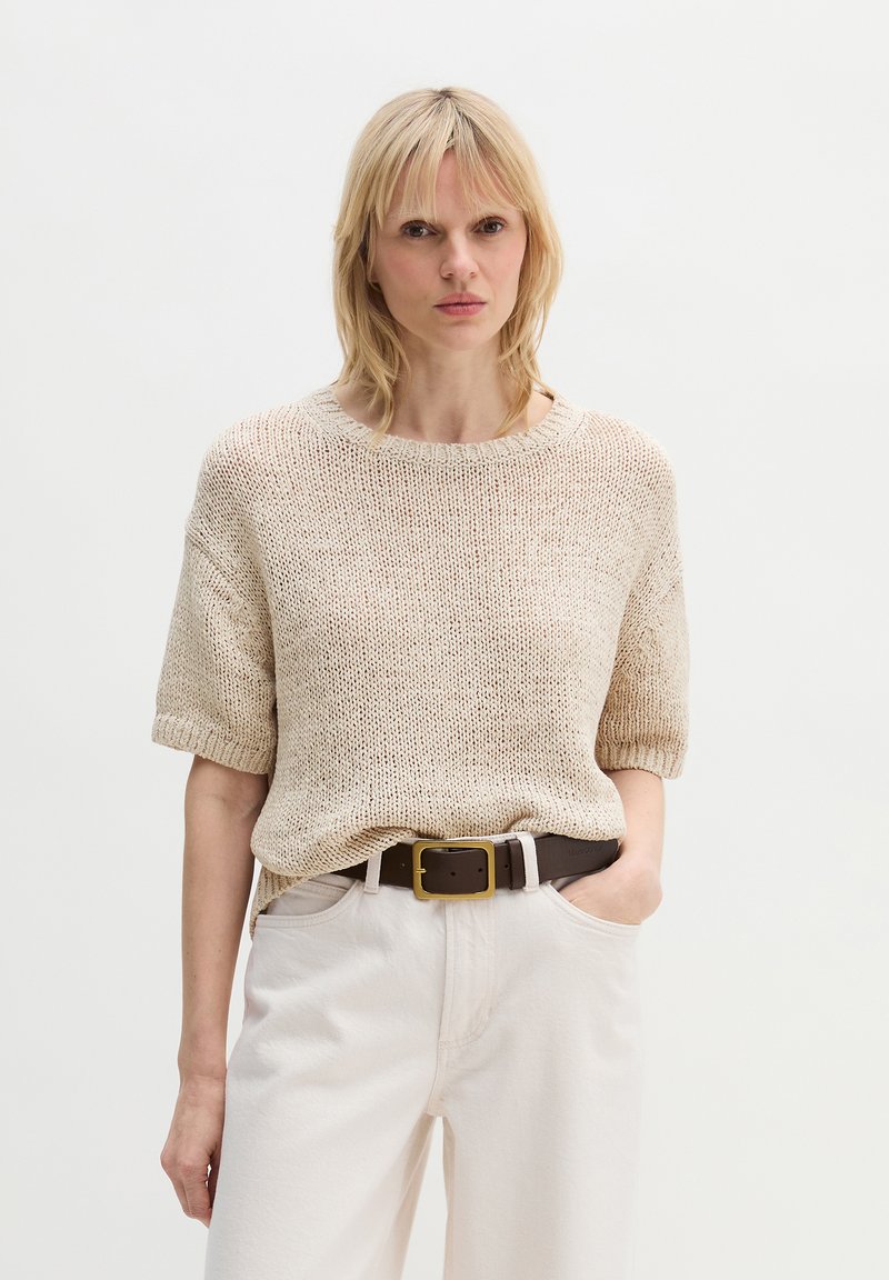 Femme blonde portant un pull beige en maille à manches courtes, rentré dans un pantalon blanc taille haute avec une ceinture marron foncé, debout devant un fond uni.