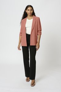 Oversized blazer v broskvové barvě s ohrnutými rukávy, v kombinaci s bílým upnutým topem a černými kalhotami. Černé sandály na opatkách dokončují celkový vzhled.