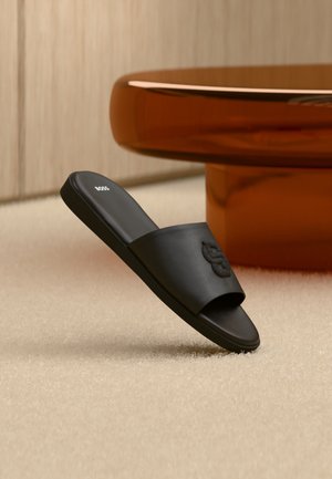 Sandalo nero tipo slide con logo in rilievo sul cinturino e scritta "BOSS" sulla soletta, posto su tappeto beige vicino alla base arrotondata di un tavolo marrone.