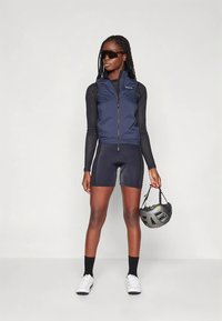 Tamsiai mėlynas dviračių liemenė su aukštu apykakliu, derinamas su juodu ilgomis rankovėmis marškinėliu ir tamsiai mėlynomis dviračių šortais. Pridedami balti sportbačiai ir pilkas šalmas.