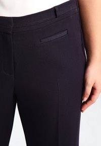 Pantalon sur mesure bleu marine avec une texture lisse, doté d'une petite poche latérale et d'une coupe droite et épurée. La finition comprend un détail de couture subtile.