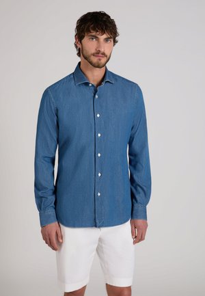 Mężczyzna o kręconych włosach, ubrany w zapinaną na guziki dżinsową koszulę i białe szorty do kolan na jednolitym tle.