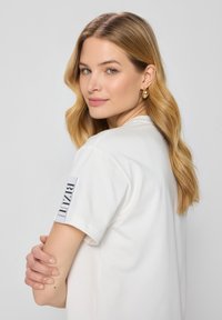 Biała bawełniana koszulka z krótkim rękawem, z czarną etykietą "BIZAU" na lewym rękawie. Modelka ma falujące blond włosy i złote kolczyki w kształcie kół.