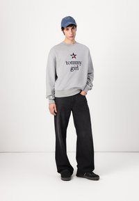 Felpa grigia con il testo "tommy girl" in blu scuro, abbinata a jeans neri a gamba larga e sneakers nere. Un cappellino blu accentua l'outfit.