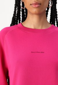 Fuksjowa bluza z zaokrąglonym dekoltem, ściągaczami przy rękawach i rękawami raglanowymi. Zawiera haftowane logo "Marc O'Polo DENIM" w dopasowanym kolorze.