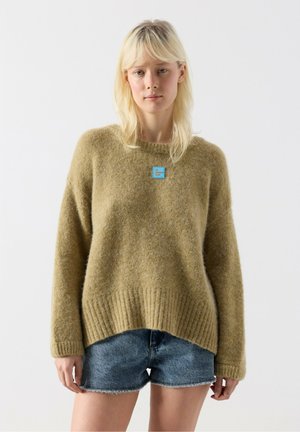 Kvinde med blondt hår iført en løs olivengrøn sweater og blå denimshorts, stående mod en ensfarvet hvid baggrund.