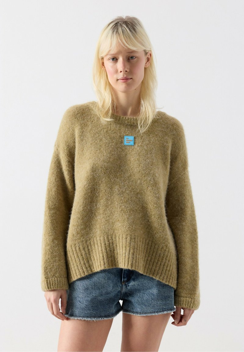 Femme aux cheveux blonds portant un pull ample vert olive et un short en jean bleu, debout devant un fond blanc uni.