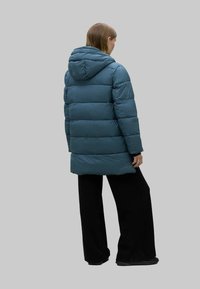 Chaqueta acolchada verde azulado con capucha, con un ajuste largo y relajado y un diseño de acolchado horizontal. Llevada sobre pantalones holgados negros.