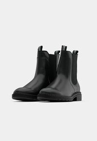 Paire de bottines en cuir noir avec des panneaux latéraux élastiques et des languettes à rayures, présentées sur un fond gris clair.