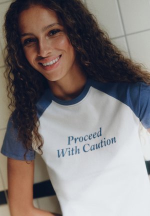 Femme souriante aux cheveux bouclés portant un t-shirt blanc et bleu avec le texte "Proceed With Caution" à l'avant.