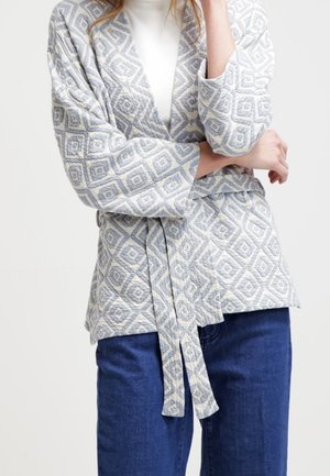 Kobieta ubrana w wzorzysty kardigan z paskiem, na białej bluzce i niebieskich dżinsach, stojąca z jedną ręką skrzyżowaną, a drugą przy twarzy.