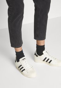 Zapatillas Adidas Superstar de cuero blanco con rayas negras, usadas con calcetines negros, combinadas con pantalones de mezclilla oscuros recortados.