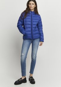 Modrá bunda typu puffer s prešívanou textúrou a predným zipsom, skombinovaná s poškodenými skinny džínsami a hrubými čiernymi čizmami.