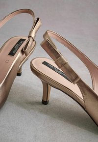 Bronze high-heeled sandals with a slingback design and decorative bow detail. Smooth metallic finish and pink insoles. Black heel accents.