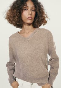 Giovane con capelli ricci che indossa un maglione beige a V e pantaloni bianchi, in piedi con le mani nelle tasche davanti a uno sfondo neutro.