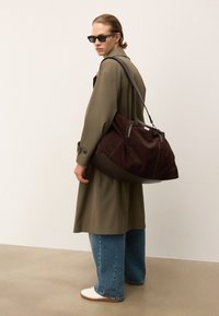 Person der bærer solbriller, olivengrøn trenchcoat, blå jeans, hvide sko, og bærer en stor mørkebrun ruskinds skuldertaske, stående på en beige gulv.