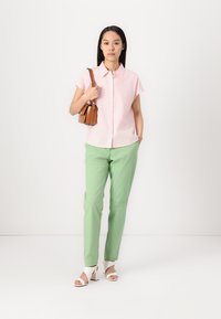 Camisa de mangas curtas às riscas rosa, calças verdes e sandálias de salto brancas. Segurando uma mala castanha. Fundo simples.