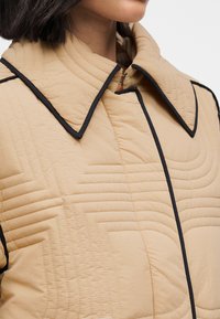 Μπεζ μπουφάν με καπιτονέ ύφασμα και δομημένο γιακά, με μαύρο σιρίτι και γραμμικά σχέδια ραφής για υφή. Απλός, σύγχρονος σχεδιασμός.