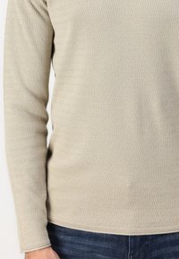 Top de manga larga beige claro que presenta una tela texturizada con un patrón de olas. El corte se ajusta al cuerpo con un dobladillo redondeado en la cintura.