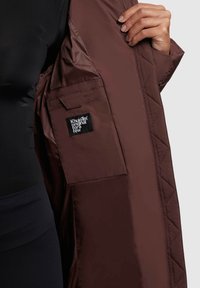 Hnědá podšívka prošívané bundy s černou nášivkou. Hladká, lesklá látka s boční kapsou a viditelným akcentním prošíváním.