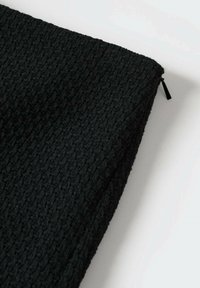 Pochette en tissu noir texturé avec une fermeture éclair dans le coin supérieur droit sur fond blanc.