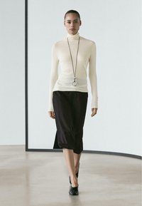 Knit turtleneck sweater in light cream, paired with a dark, sleek skirt. Model wears a silver pendant and black, pointed-toe shoes.