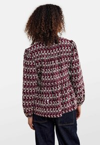 Blusa marrón con mangas largas abullonadas, con un patrón floral blanco. Tejido texturizado con detalles fruncidos en la parte posterior. Ajuste casual.