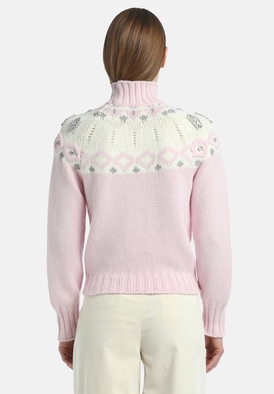 Kobieta w różowo-białym dzianinowym swetrze z wysokim kołnierzem i ozdobnymi koralikami na górnej części pleców i ramionach.