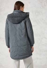 Manteau d'hiver long gris foncé matelassé avec une capuche en polaire grise texturée, des poignets élastiques et un ourlet légèrement arrondi. Tissu lisse avec des coutures minimales.