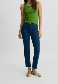 Zelený žebrovaný top bez rukávů s logem, kombinovaný s modrými džíny s vysokým pasem a rovnými nohavicemi, a metalickými zlatými balerínami.