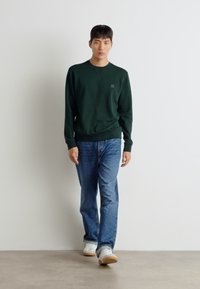 Donkergroen sweatshirt met een ronde hals en ribgebreide manchetten, gecombineerd met lichtblauwe jeans en witte sneakers, staande op een neutrale achtergrond.