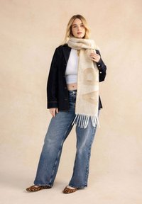 Beige wollen sjaal met franjes, voorzien van een subtiel patroon. G gedragen met een spijkerjack en wijde jeans tegen een neutrale achtergrond.