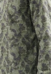 Groen camouflage stof met een gestructureerd patroon van donkere bladvormen, wat een gelaagd, gemengd uiterlijk creëert.