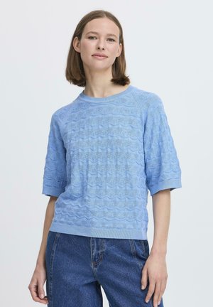 Giovane donna con capelli castano chiaro di media lunghezza, che indossa un maglione azzurro a maniche corte con trama e jeans blu scuro, in piedi contro uno sfondo bianco.